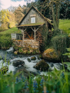 Rustic wooden cottage above a flowing stream with rocks and greenery