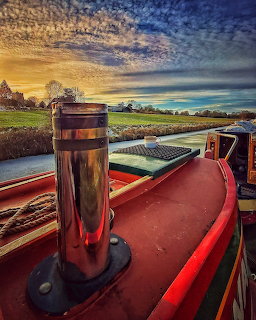 Front of a red narrowboat on a canal beneath a dramatic sunset sky