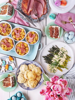 Overhead Easter meal with ham, asparagus, biscuits, dyed eggs, desserts, and pink flowers on a table