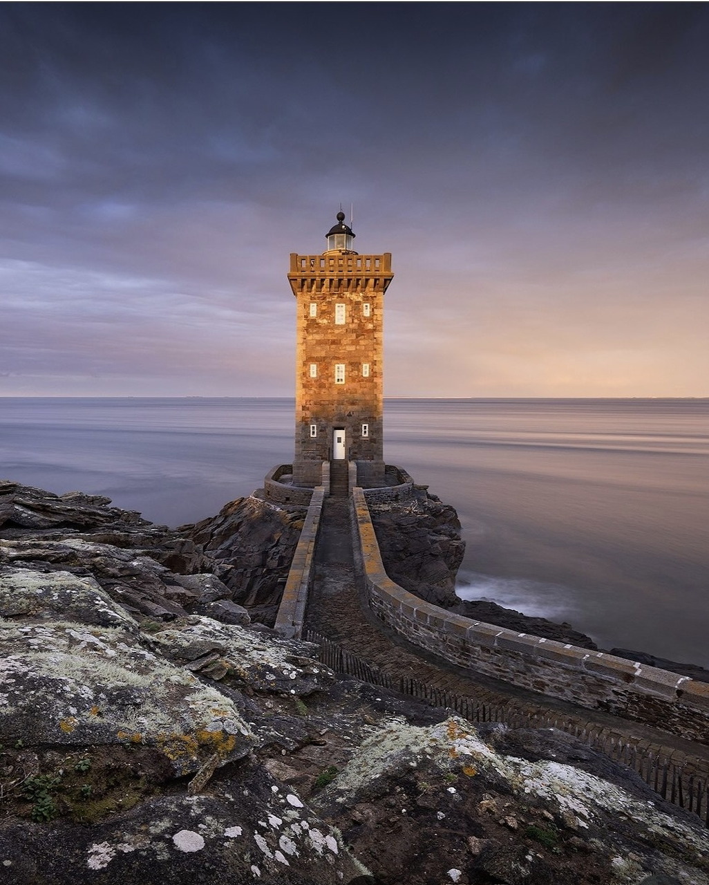 Illuminated stone lighthouse on rocky coastline at dusk with calm ocean and cloudy sky