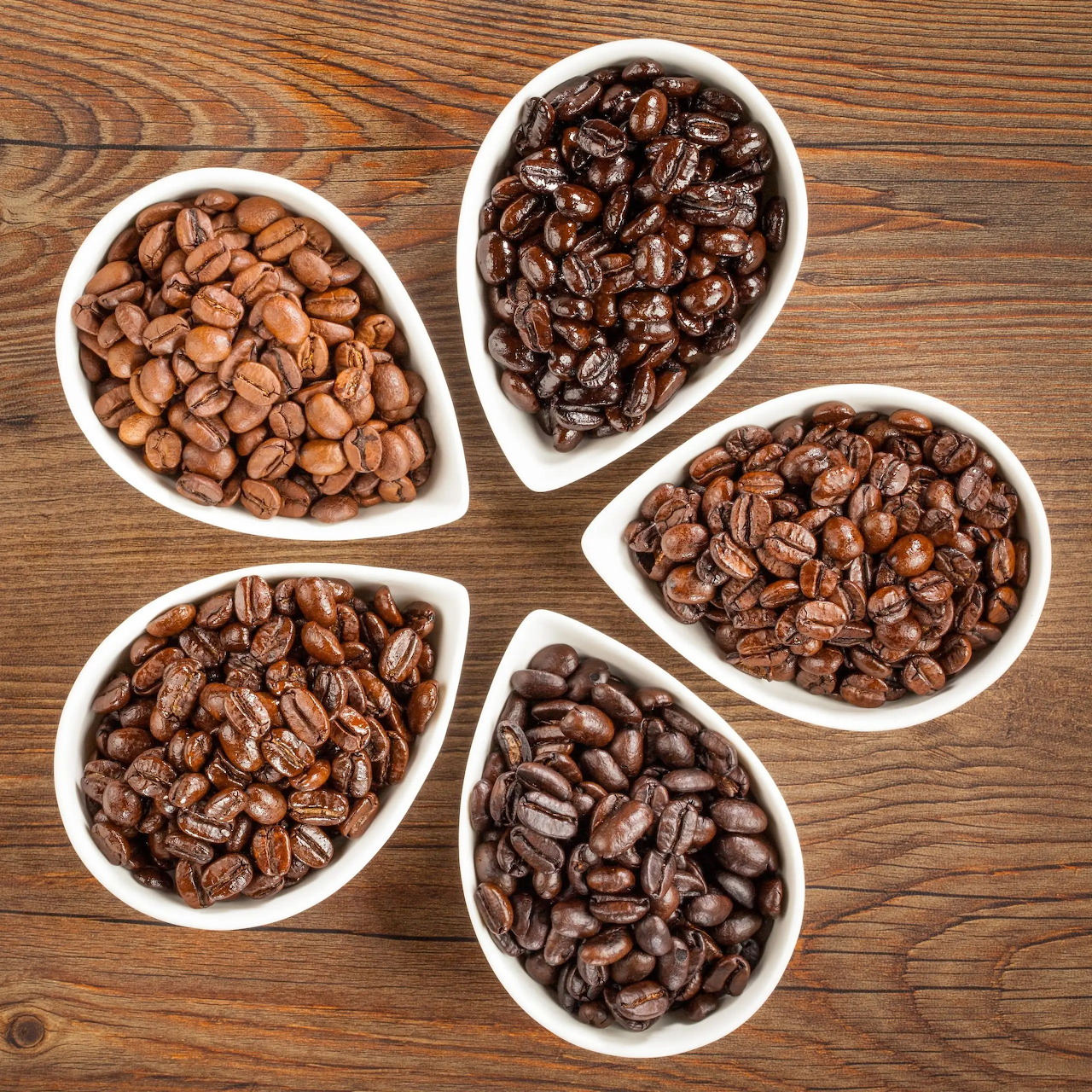 Five white bowls containing various roasted coffee beans on wood surface