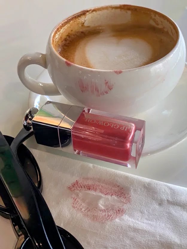 Cappuccino cup with foam, pink lip gloss, lipstick kiss stain on cloth, and sunglasses on white table