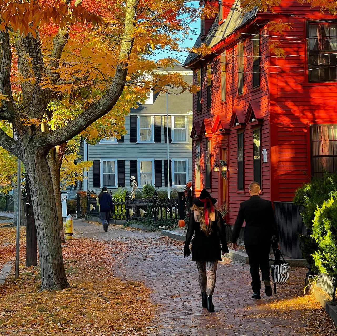 People in costumes walking on brick street past colonial houses with autumn trees in Salem during Halloween