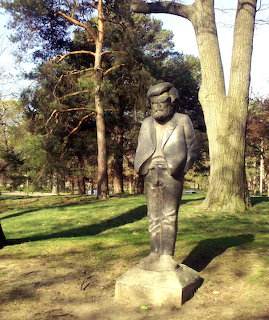 Stone sculpture of a hippie figure in a park with tall trees and green grass