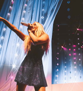 Blonde woman in black sparkly dress singing on stage with blue curtains and crystal decorations