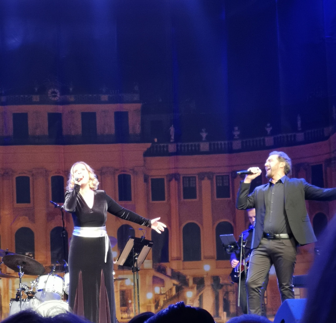 Two opera singers in formal attire performing on stage at night with illuminated historic building behind them
