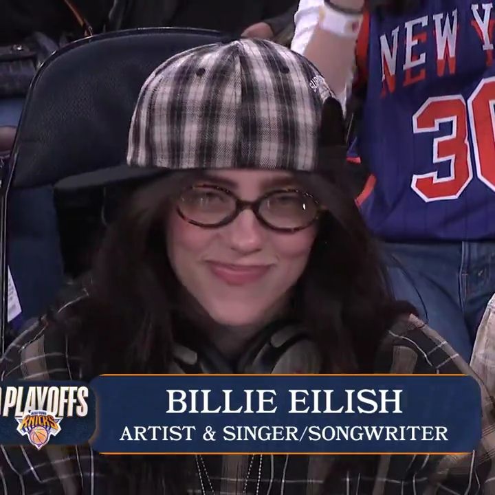 Billie Eilish in glasses and a plaid cap with a broadcast caption at a basketball game