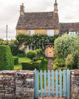 Stone cottage with climbing plants, green garden, and blue wooden gate in front