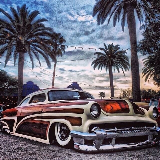 Vintage low-rider car parked beneath palm trees under a cloudy evening sky