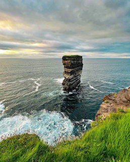 Tall rock sea stack jutting from ocean beside grassy clifftop under cloudy sky at Downpatrick Head, Ireland