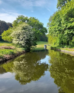 Calm canal with tree reflections, grassy banks, and a small boat in the distance