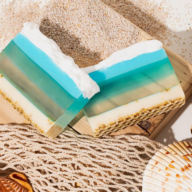 Two handmade soap bars with layered turquoise, white and tan colors on woven mat with sand and shells