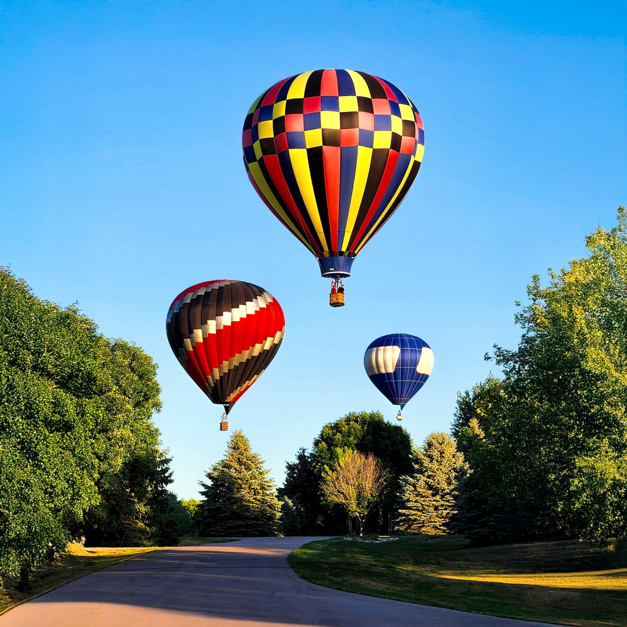 Three colorful hot air balloons flying over a tree-lined road beneath a blue sky