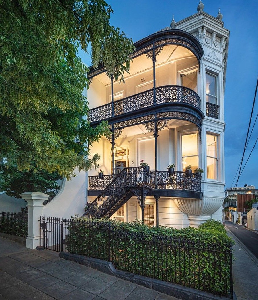 Victorian terrace house with ornate balconies illuminated at dusk with green trees in foreground