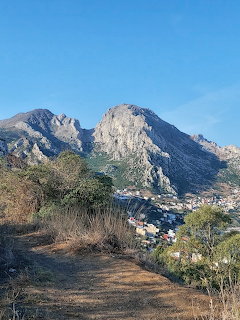 Rocky mountain towering over a small village surrounded by green trees and vegetation under blue sky