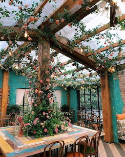 Cafe interior with glass roof pergola covered in pink roses, wooden beams, turquoise walls, and rustic dining tables