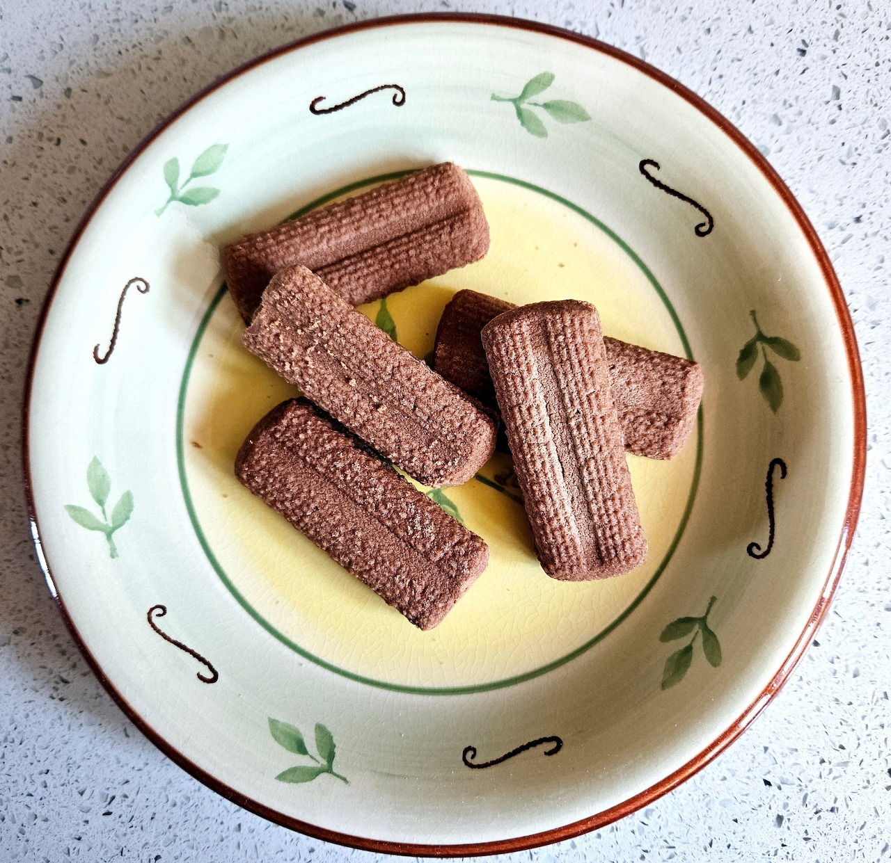 Four chocolate wafer roll cookies on a yellow-centered plate with green leaf design