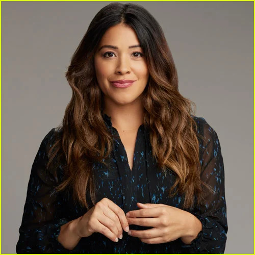 Smiling woman with long wavy brown hair in a dark blouse, posed against a plain studio background