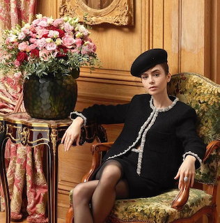 Lily Collins seated in a floral chair wearing a black dress and beret beside pink flowers
