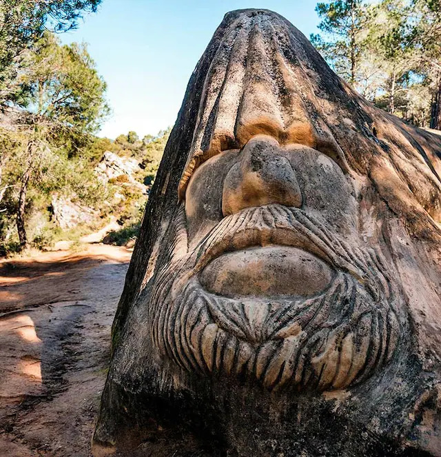 Carved stone face with beard and prominent features sculpted into large rock formation in forest setting