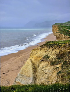 Tall eroded cliff beside empty beach with ocean waves and distant headland under cloudy sky
