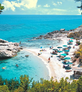 Aerial view of Greek beach cove with turquoise water, white sand, rocks, and colorful umbrellas
