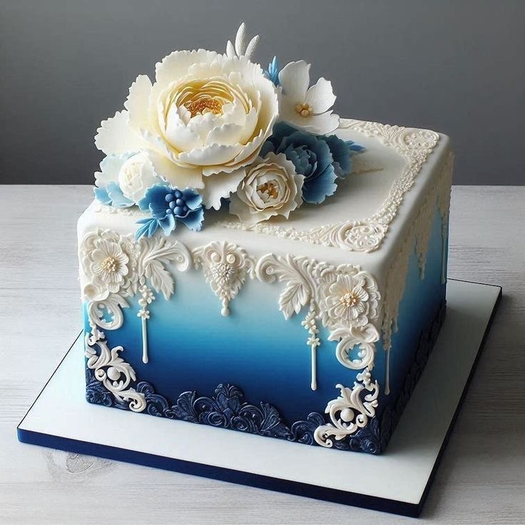 Square cake with blue ombre frosting, ornate white details, and cream sugar flowers