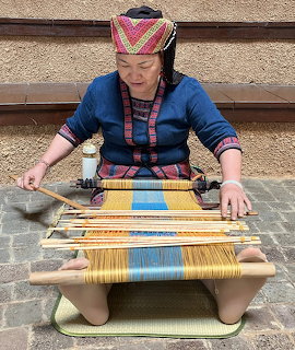Woman in traditional clothing weaving colorful fabric on a wooden loom while seated outdoors