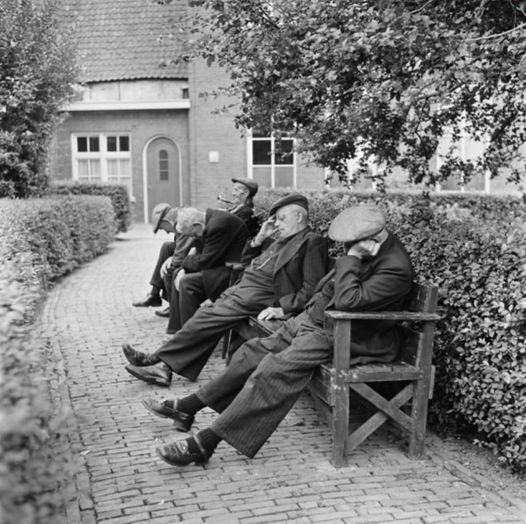 Black-and-white image of elderly men sitting on a bench beside a cobblestone path near a house