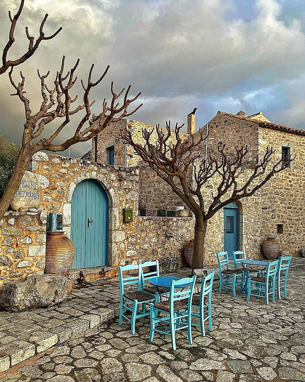 Stone village courtyard with blue doors, turquoise chairs, and leafless trees