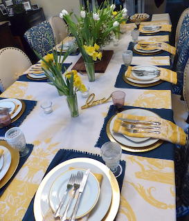 Formal dining table set with yellow placemats, white plates, blue napkins, and yellow floral centerpieces
