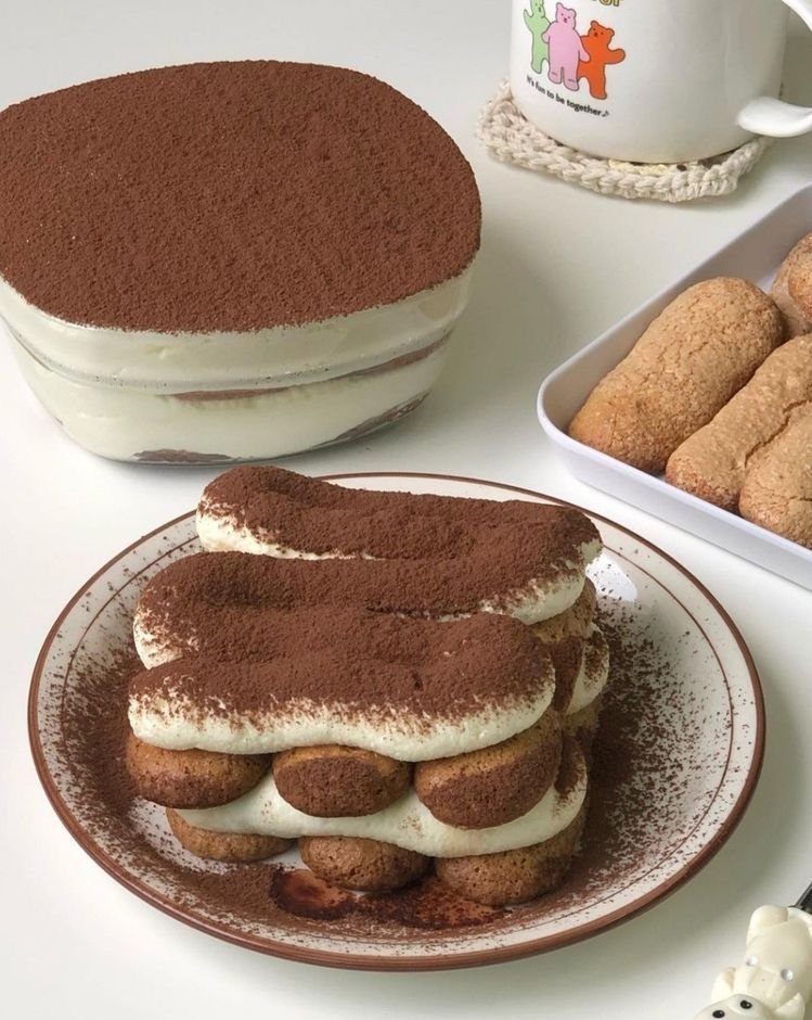 Plated tiramisu dusted with cocoa beside a dish of tiramisu and ladyfinger biscuits