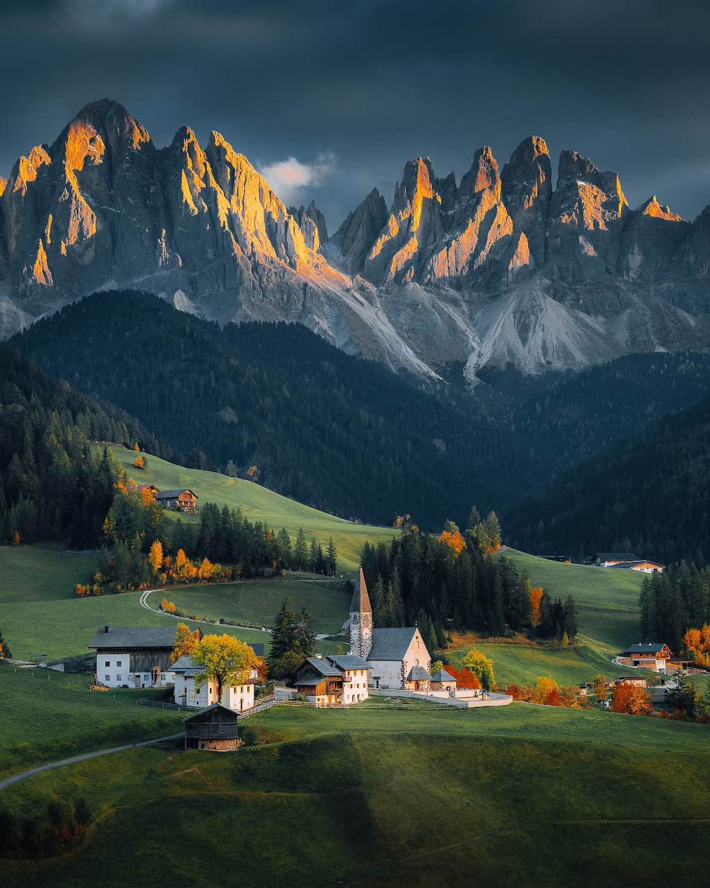 Alpine village with church in green valley below dramatic Dolomite mountain peaks in autumn