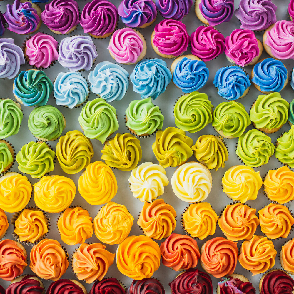 Grid of cupcakes with rainbow-colored swirl frosting arranged in horizontal rows by color