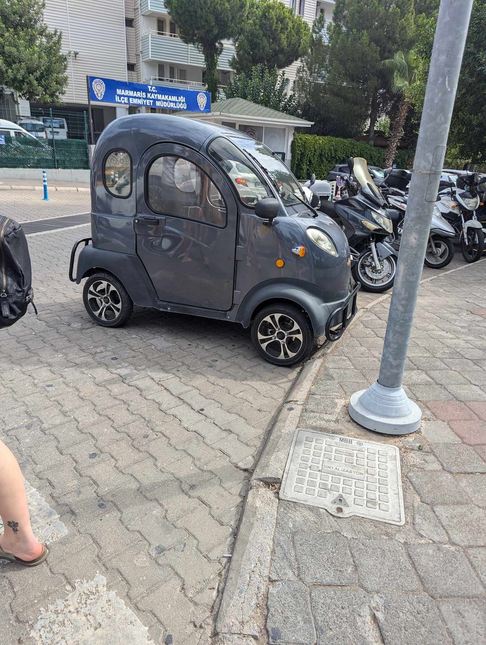 Tiny dark gray micro electric car parked on cobblestone street with scooters in background