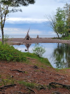River mouth with driftwood teepee structures on a sandy lakeshore surrounded by trees