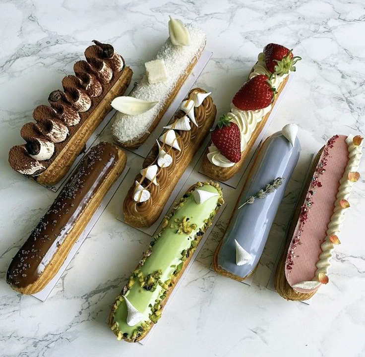Six colorful gourmet éclairs with various toppings on marble surface