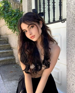 Young woman with long dark wavy hair sitting outdoors in a black top near a white door