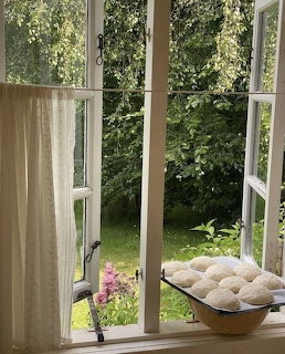 Open white cottage window with linen curtain, bread dough on sill, green garden and pink flowers outside