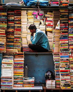 Man sitting in a small street bookshop surrounded by dense stacks of colorful books