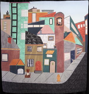 Fabric quilt artwork showing colorful geometric houses in a village arrangement with patchwork patterns and small cat