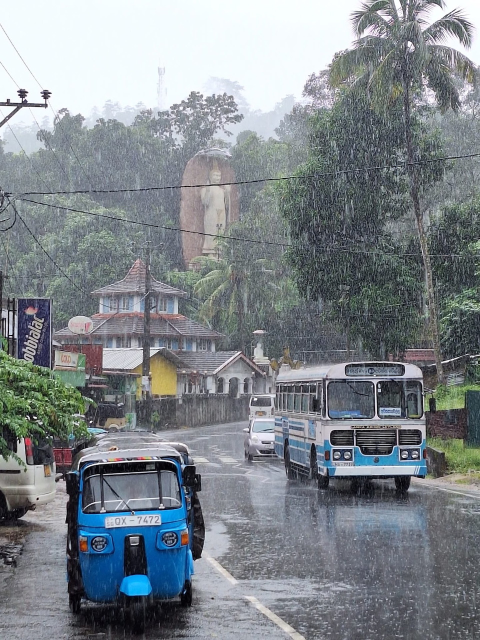 Rainy street in Sri Lanka with tuk-tuk, bus, and hillside Buddha statue amid green trees
