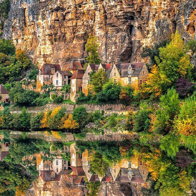 Village houses below a rocky cliff with autumn trees reflected in calm water
