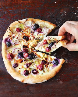 Hand lifting slice of white pizza topped with grapes and cheese on dark rustic table
