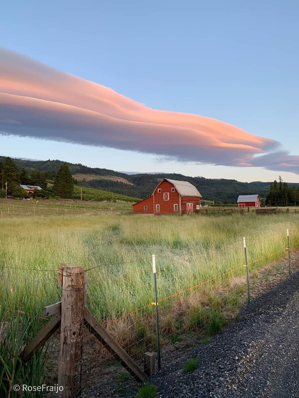 Red barn in a grassy field with a dramatic lenticular cloud glowing pink at sunset and forested hills behind