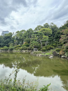 Pond with tree reflections, rocks, and greenery under a cloudy sky in a landscaped garden