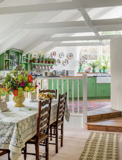 Rustic kitchen with green cabinets, white beamed ceiling, dining table with flowers, and decorative wall plates