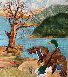 Fabric quilt art showing a mallard duck spreading its wings beside a bare tree near a lake with green hills