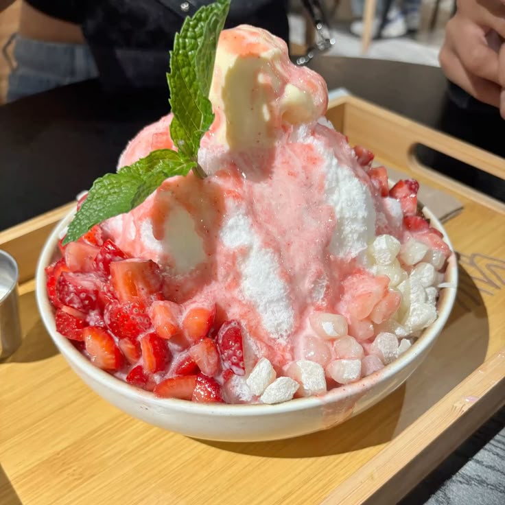 Tall mound of strawberry bingsu shaved ice with cream, fruit, and mint leaf in white bowl