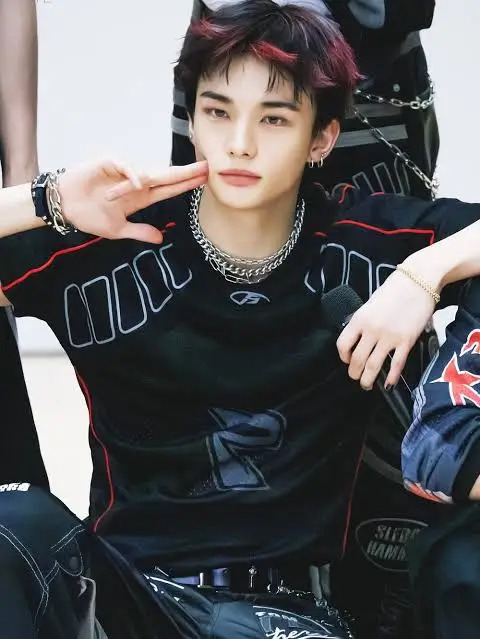 Hyunjin from Stray Kids sitting and posing with a peace sign, wearing black outfit with red hair